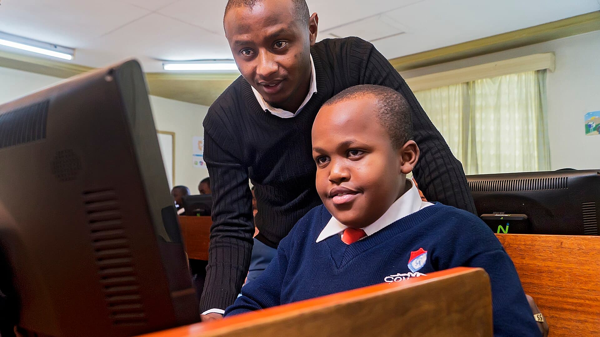 A teacher guides a smiling pupil working at a desktop computer